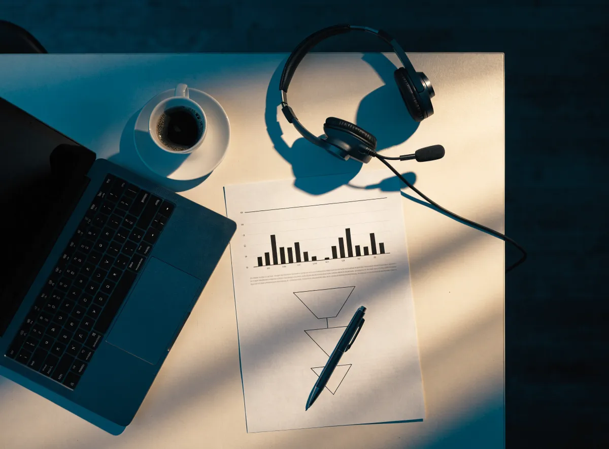 Vista cenital del informe semanal impreso de una campaña de telemarketing, con gráfico de barras y embudo, junto a unos auriculares, un portátil y un café